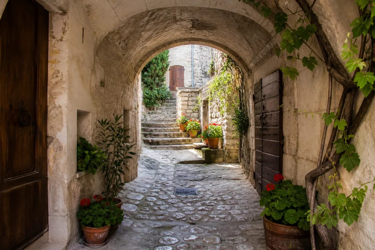 Ruelle voûtée en pierre calcaire d’un village ardéchois perché, escalier ancien et volets de bois sous une lumière douce